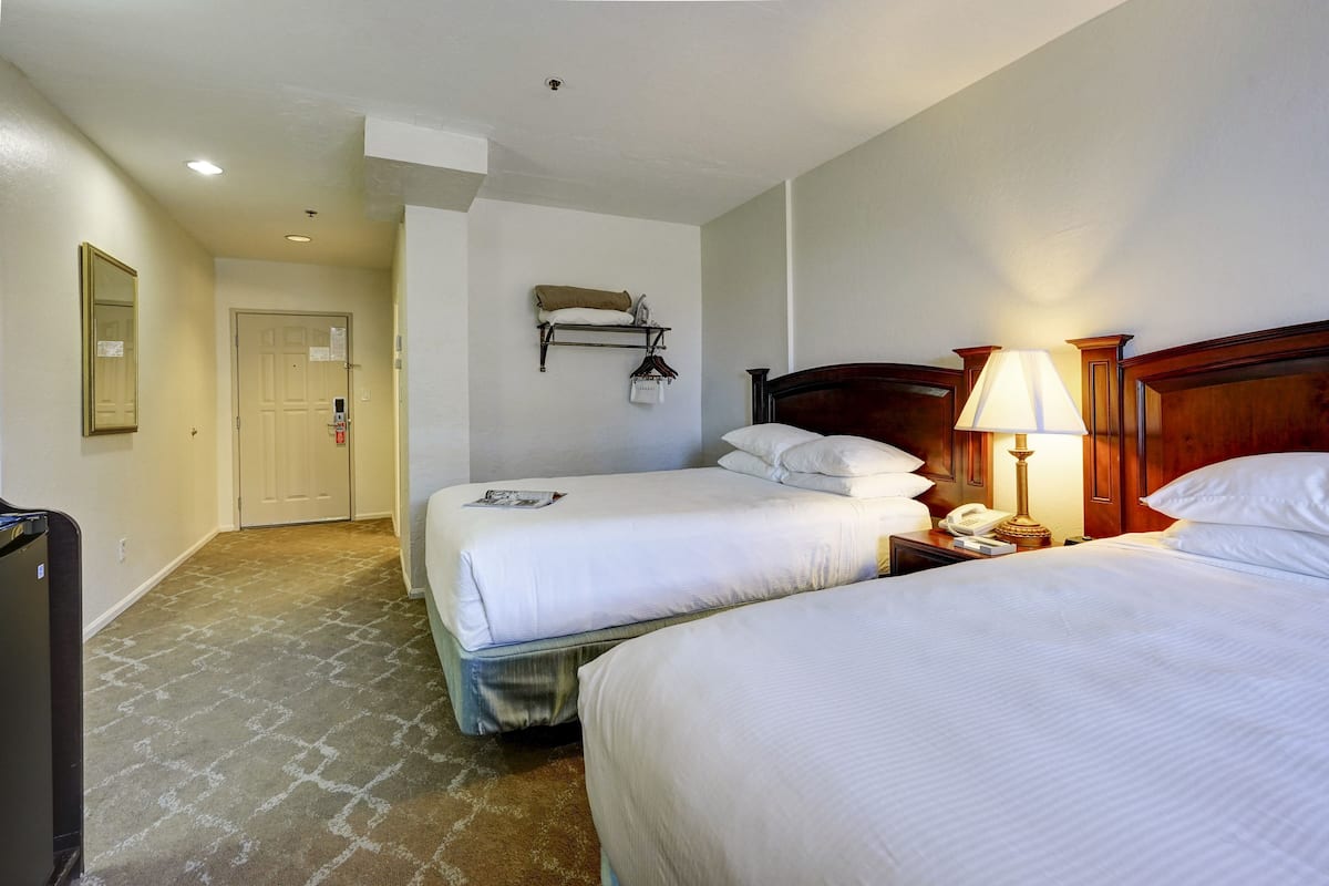 Ein Hotelzimmer mit zwei Betten, einem hölzernen Kopfteil, einem Nachttisch mit Lampe, einem Wandregal mit Kissen und Kleiderhaken sowie einer Tür mit Lichtschalter.