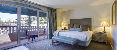 A hotel room with a large bed, two bedside lamps, a bench, a sliding glass door to an outdoor balcony, and a nightstand with a lamp.