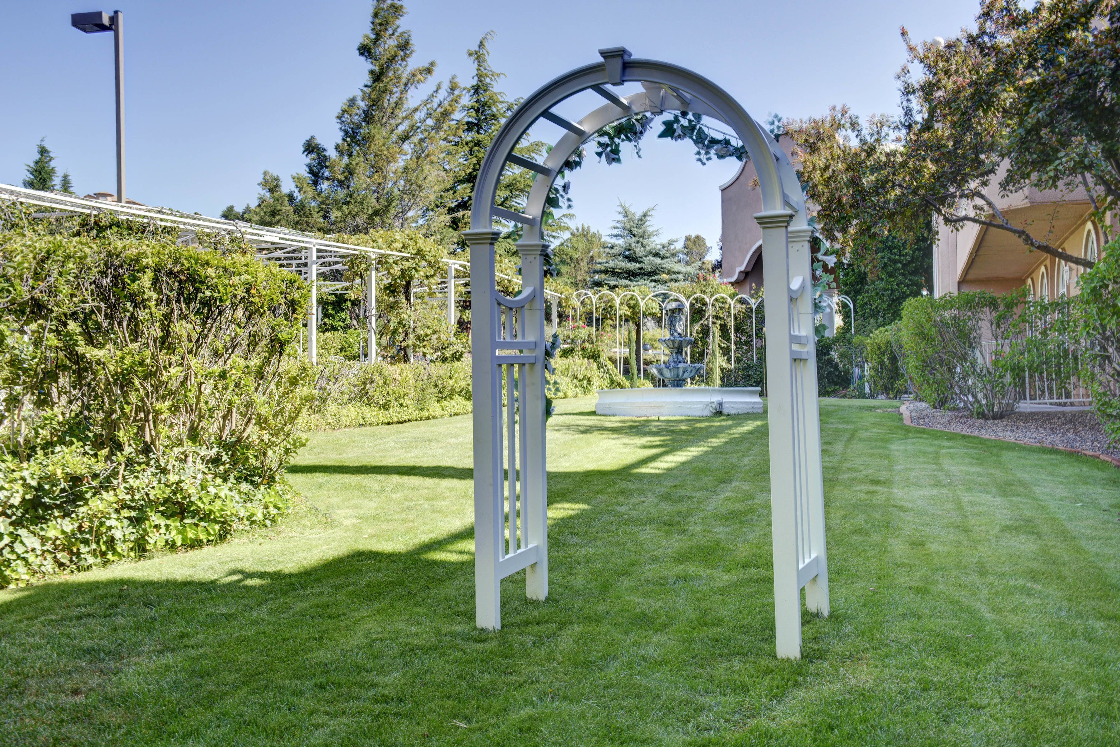 A white arched structure in a well-maintained garden with a clear sky.