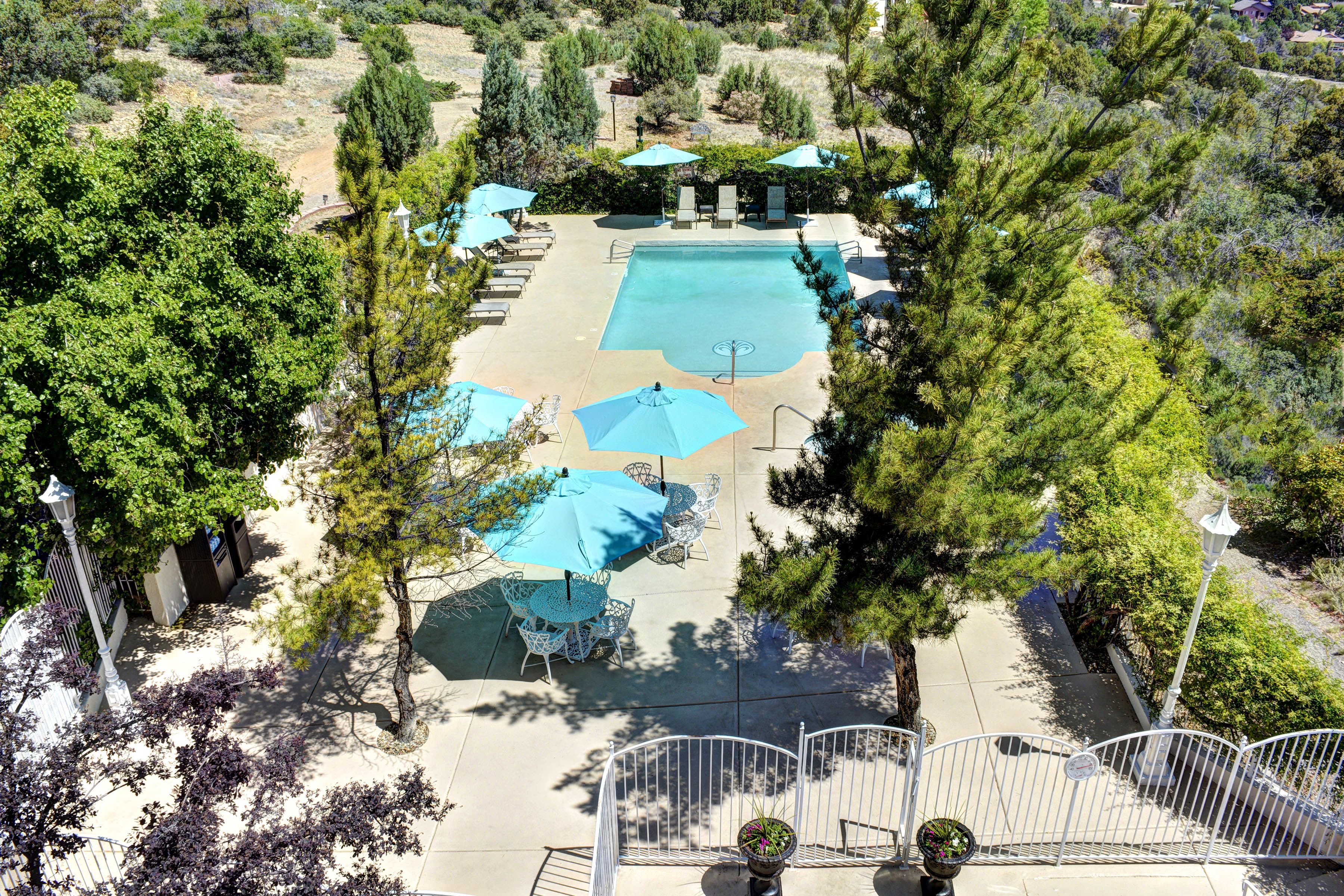 An aerial view of a resort with a central swimming pool, surrounded by lounge chairs and umbrellas, with trees and a staircase leading to upper levels.
