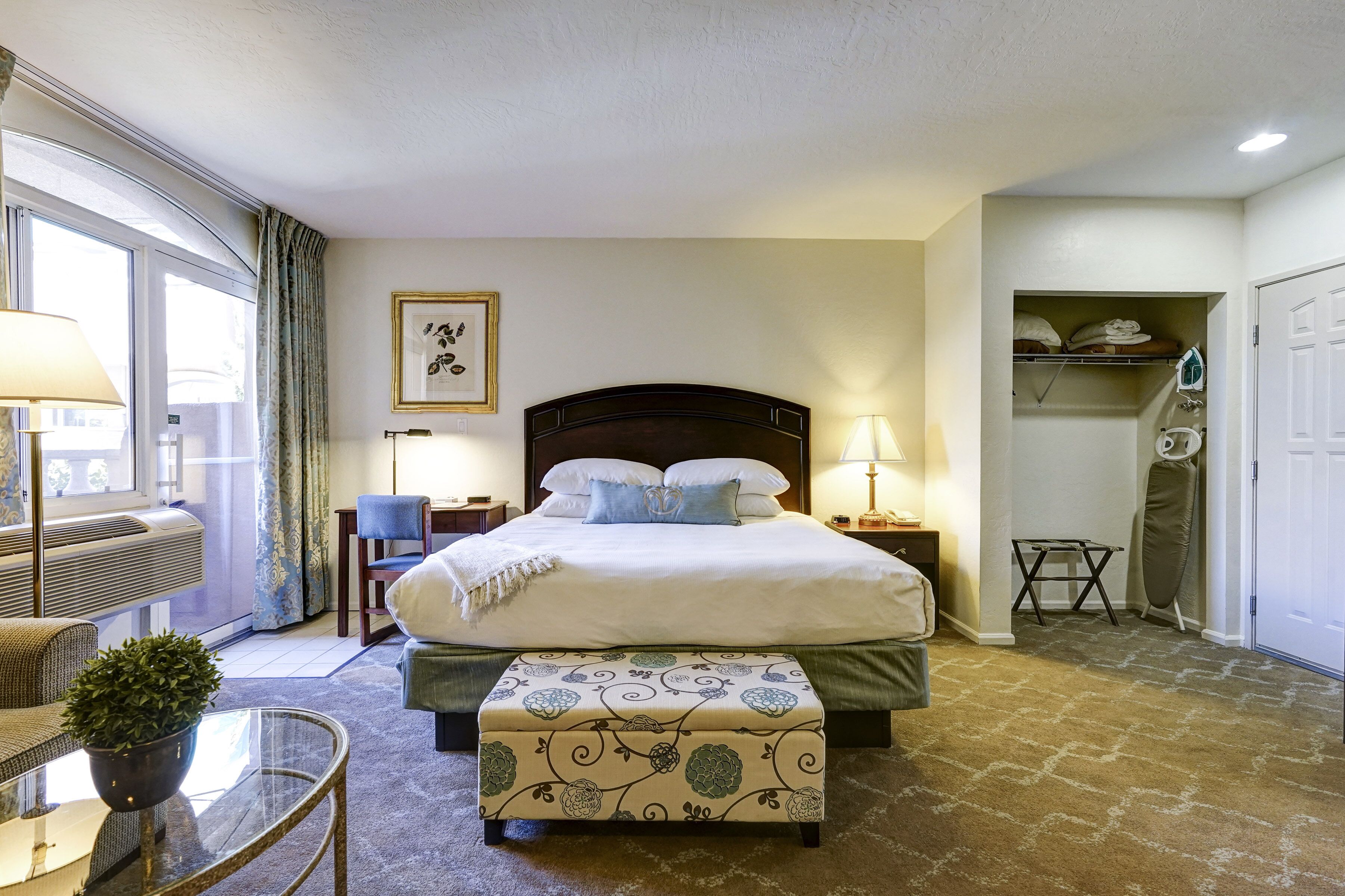 A hotel room with a large bed, a bedside table, a chair, a small table with a plant, and a view of the outside through a window.