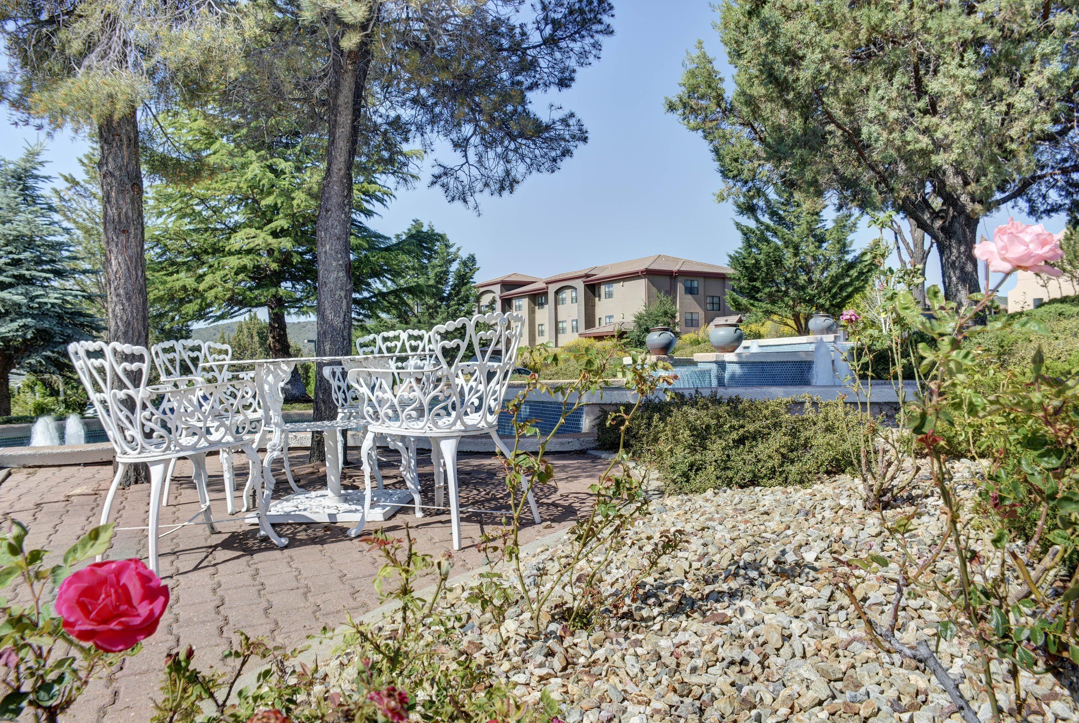 A patio area with white metal chairs and tables, surrounded by trees and a building in the background.