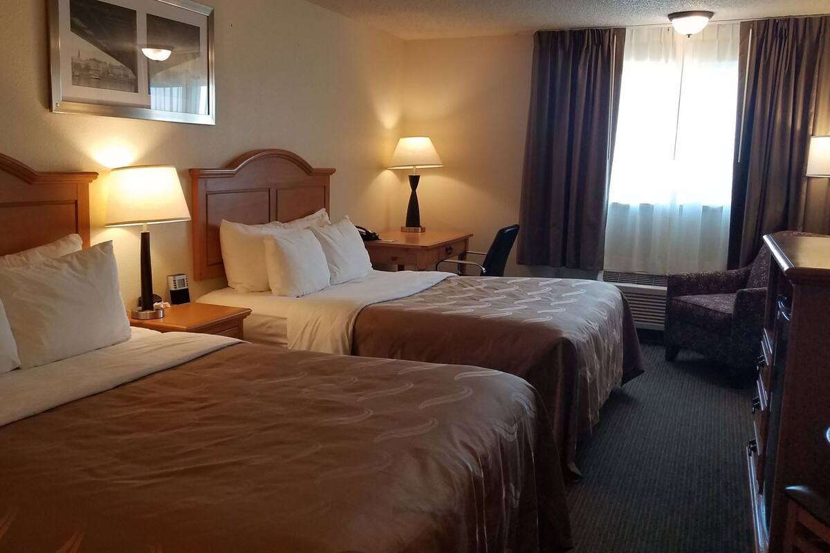 A hotel room with two beds, a desk, a chair, and a window with curtains.