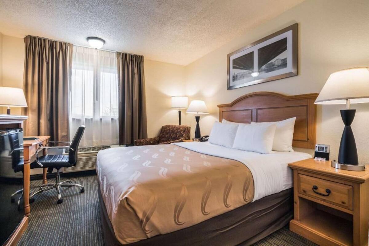 A hotel room with a bed, two bedside tables with lamps, a desk with a chair, a chair, a window with curtains, and a framed picture on the wall.