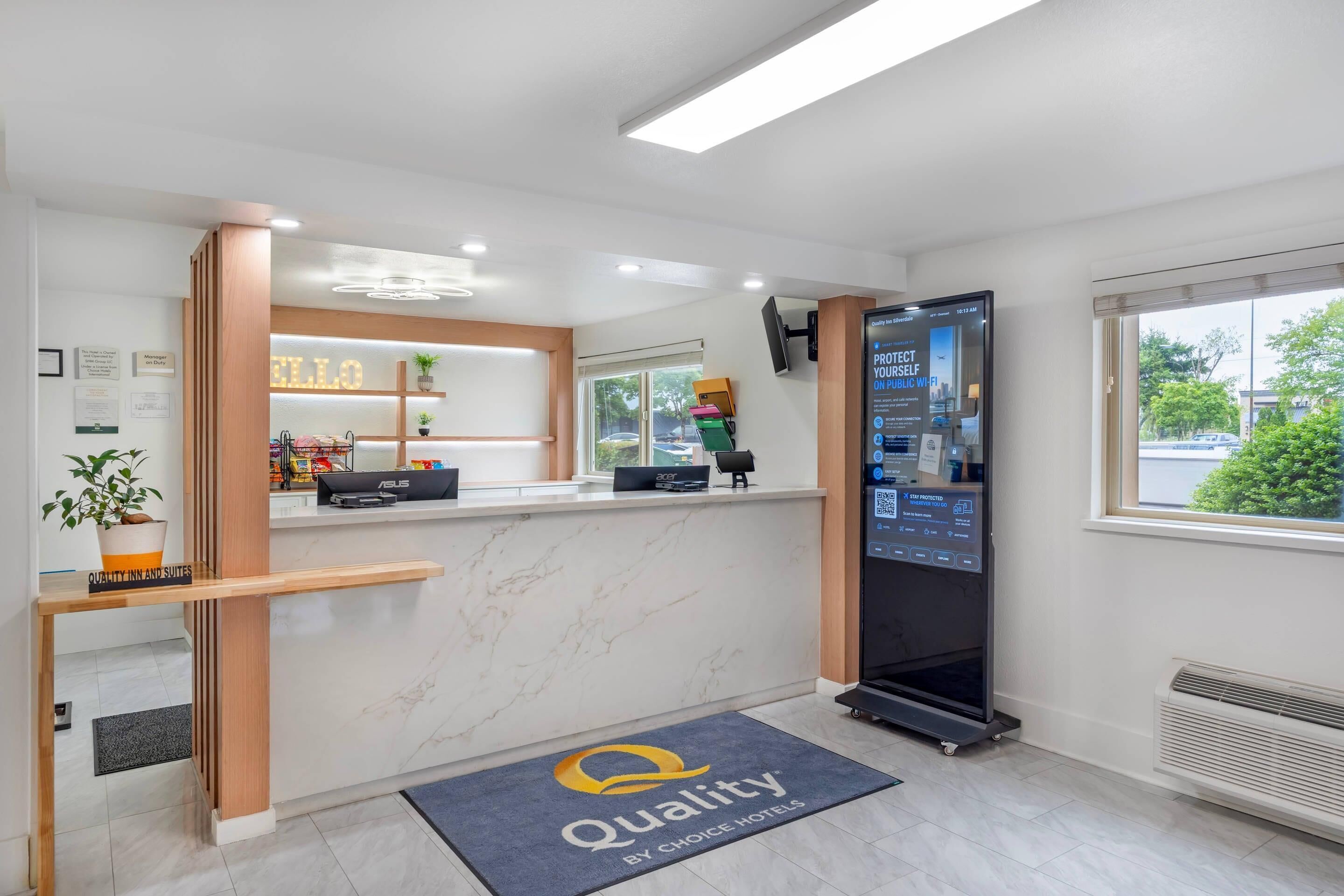 A hotel reception area with a digital information board, a reception desk, and a 'Quality' doormat.