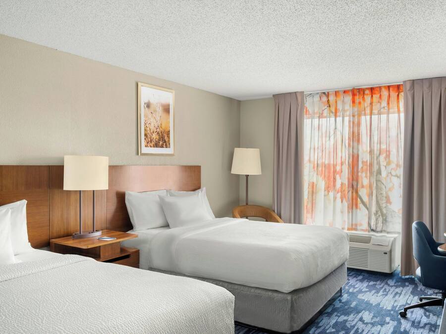 Una habitación de hotel con dos camas, un escritorio, una silla, una lámpara y un ventanal con cortinas.