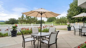 An outdoor patio area with a round table and chairs, a beige umbrella, and potted plants.