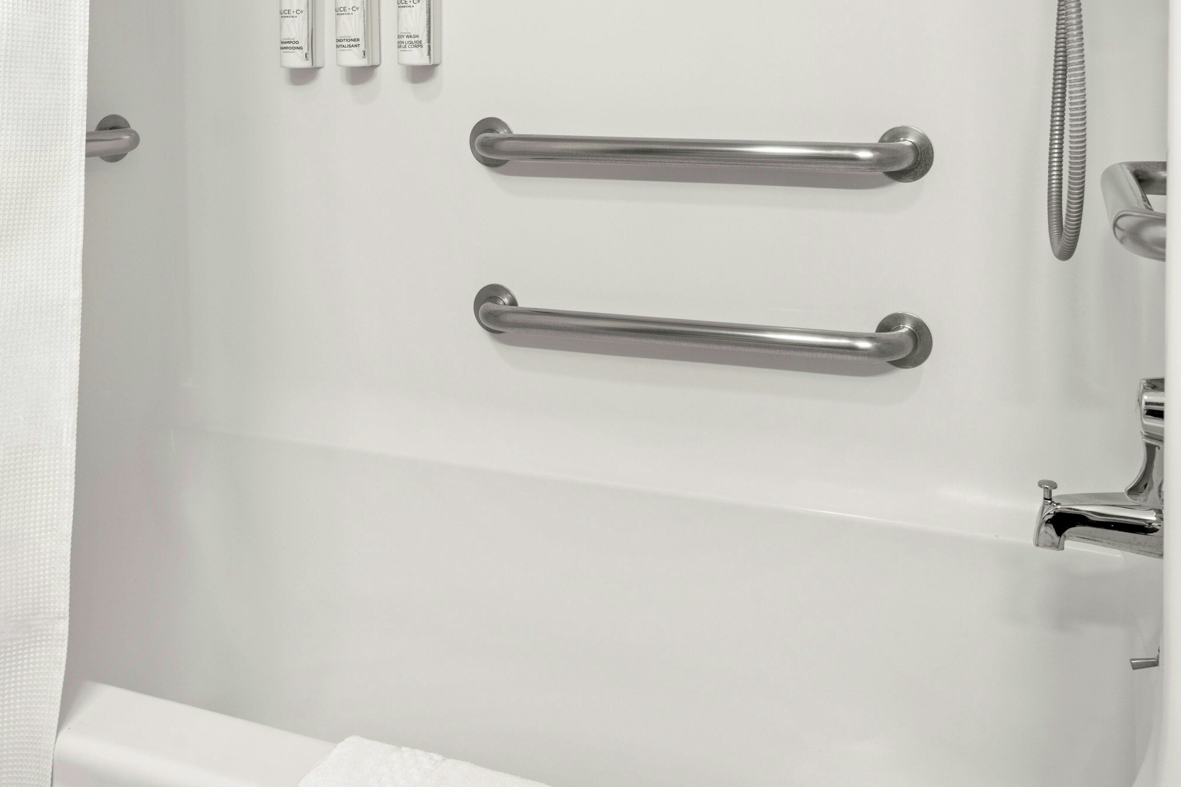Une baignoire blanche équipée de deux barres d’appui métalliques, d’un pommeau de douche et d’un robinet.
