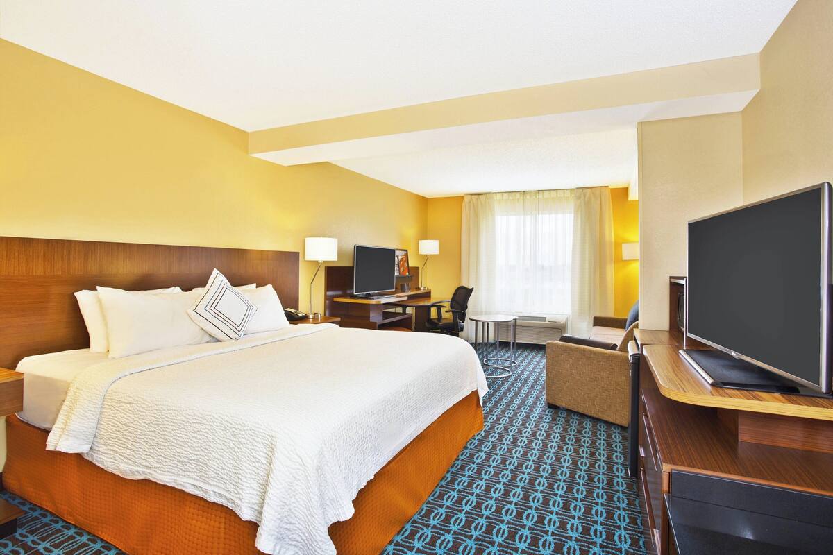 Ein Hotelzimmer mit einem großen Bett, einem Schreibtisch mit Computer, einem Fernseher und einer Sitzecke mit Sessel und Tisch.