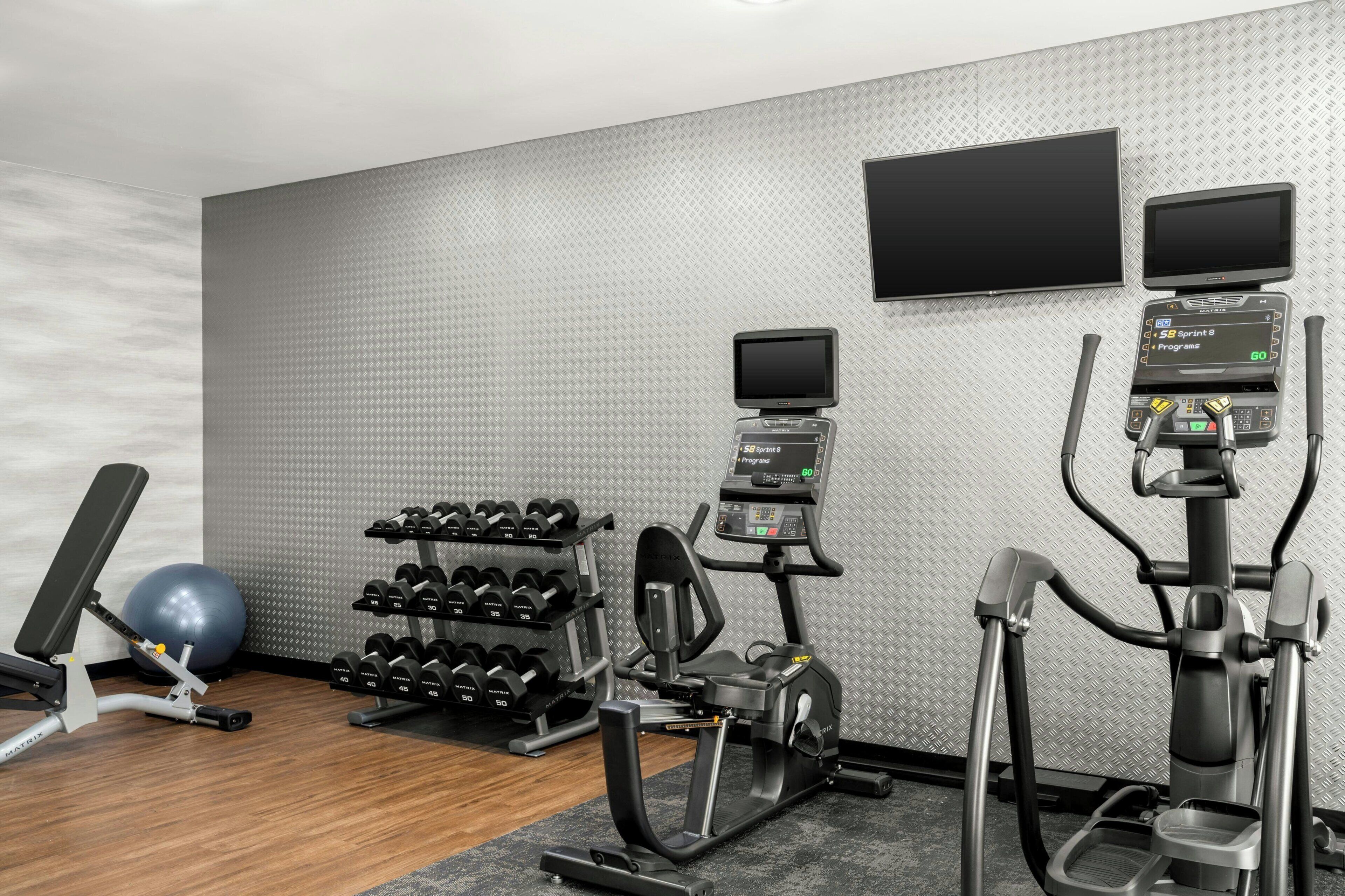 A gym with treadmills, a stationary bike, a rowing machine, a wall-mounted TV, and a rack of dumbbells.