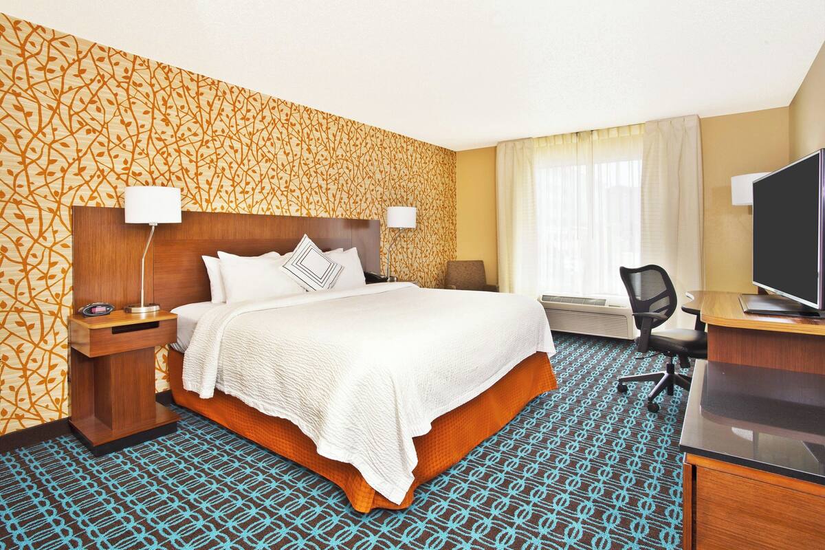 Ein Hotelzimmer mit einem großen Bett, einem Schreibtisch mit Fernseher, einem Sessel und einer Schreibtischlampe.