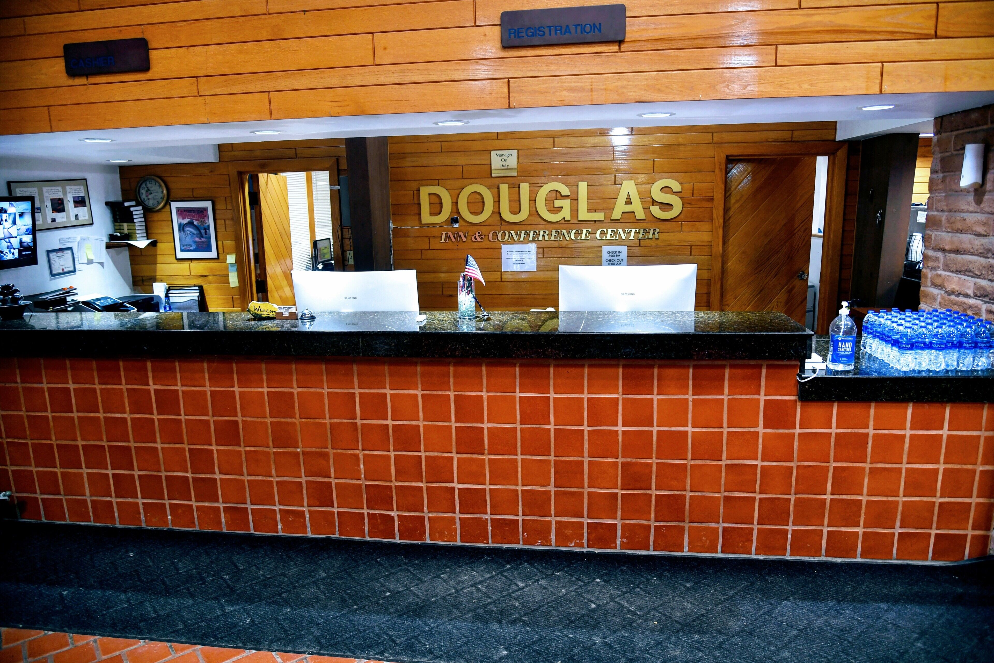 Un comptoir d'accueil à l'hôtel Douglas Inn & Conference Center, avec un panneau en bois en arrière-plan, une horloge et un panneau d'inscription.