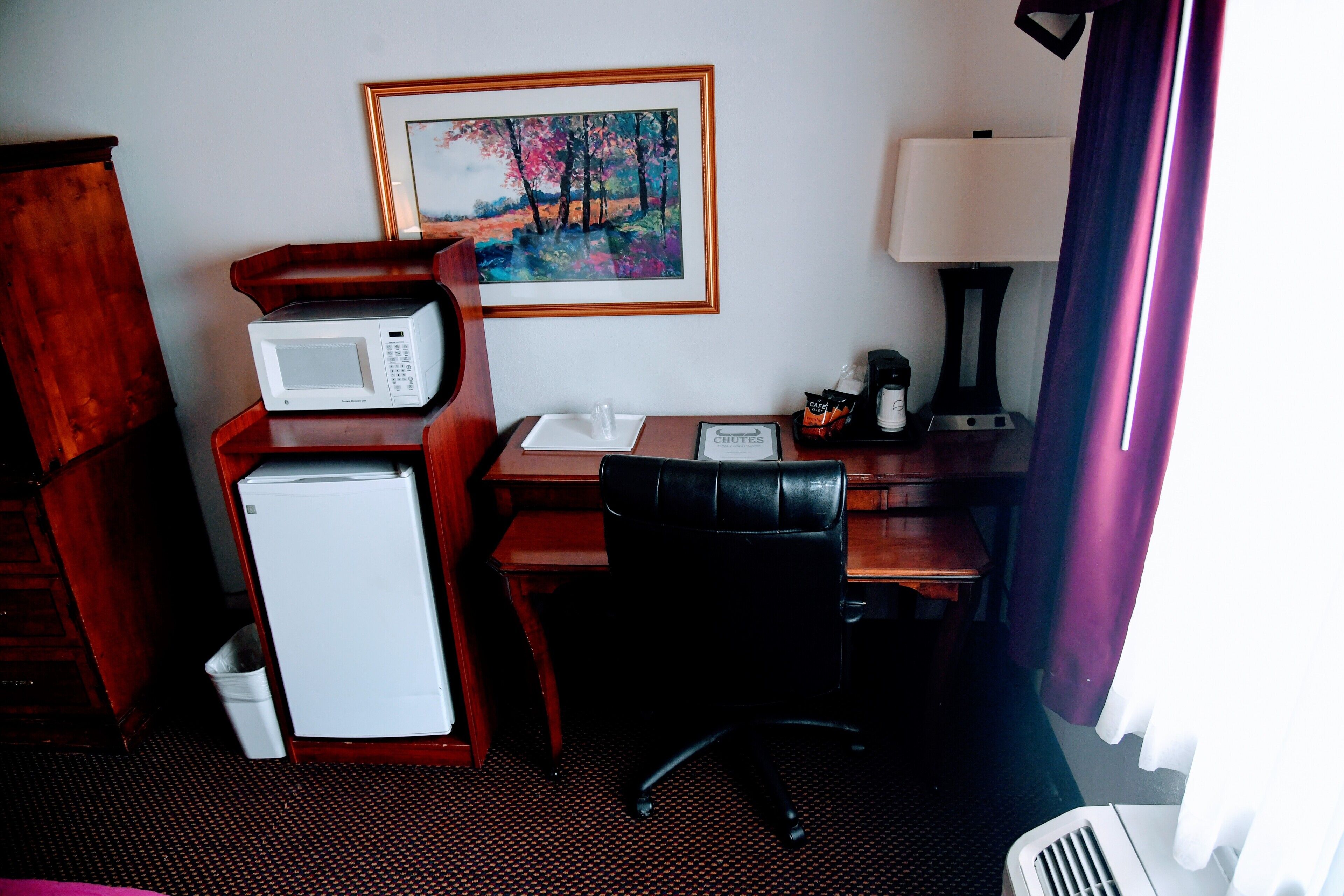 A hotel room with a wooden desk, a white refrigerator, a black office chair, a framed painting on the wall, and a lamp.