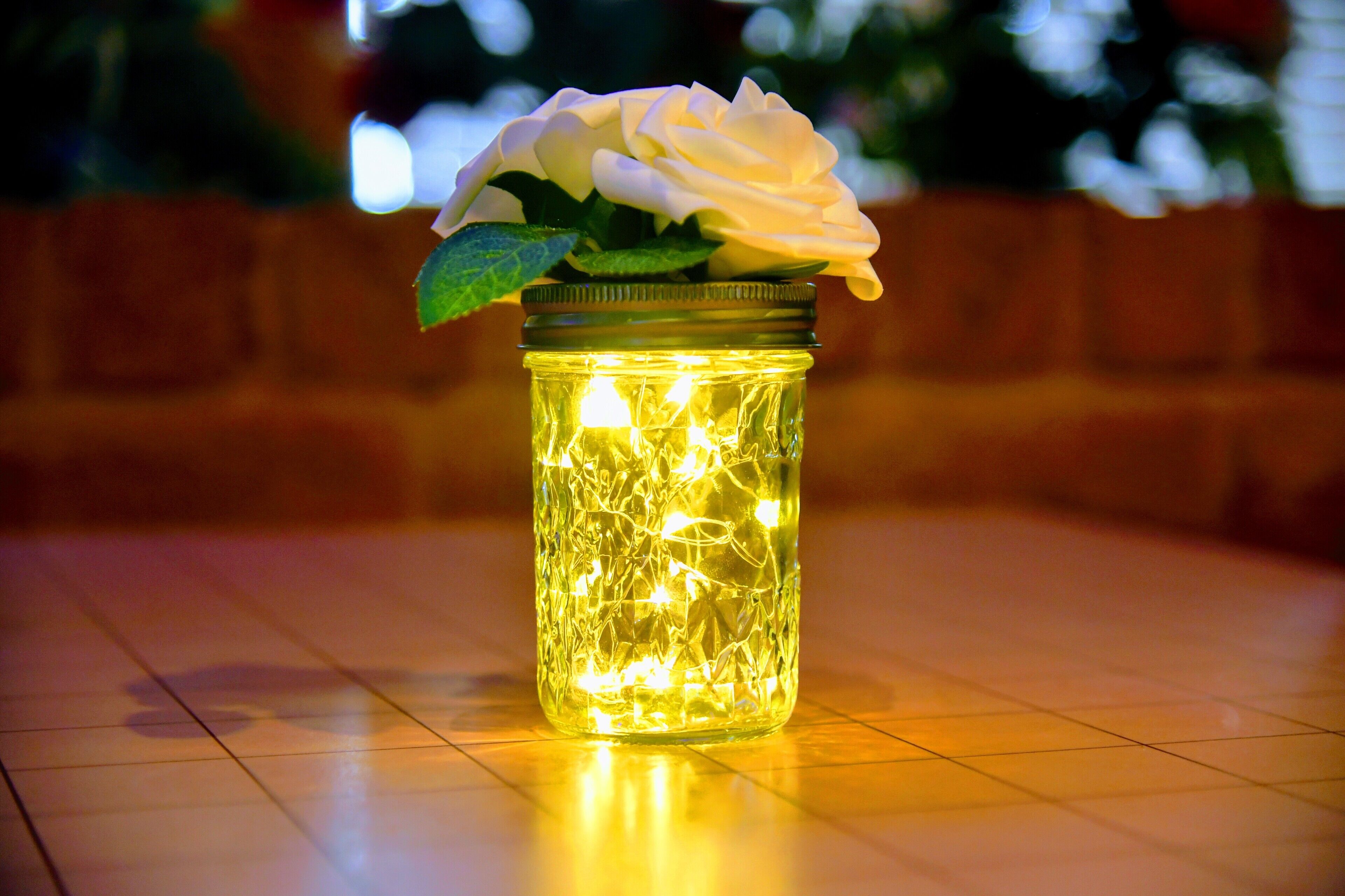 A glass jar with a white rose and string lights on a tiled floor.