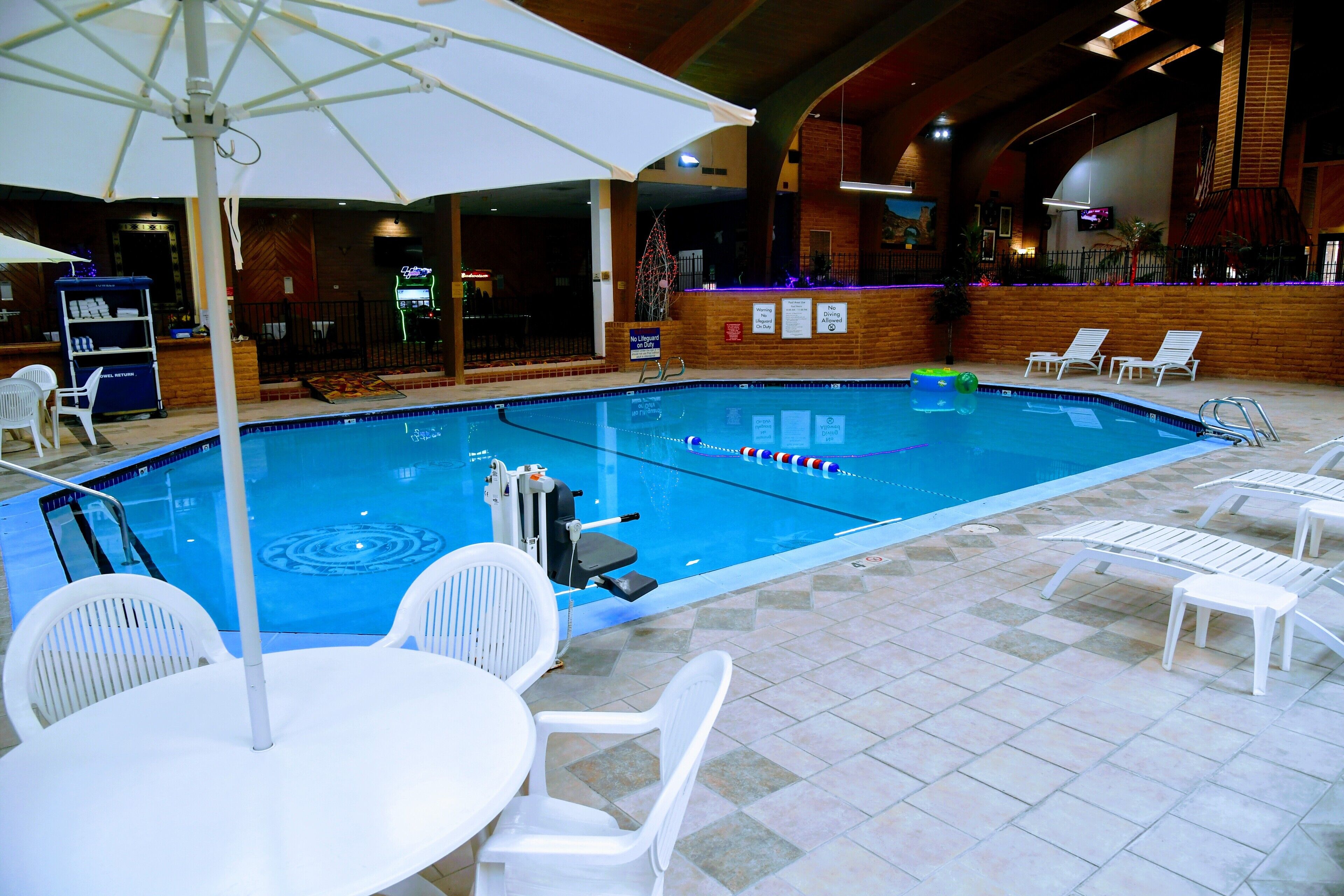 An indoor swimming pool with a tiled floor, white lounge chairs, and a bar area with a neon sign.