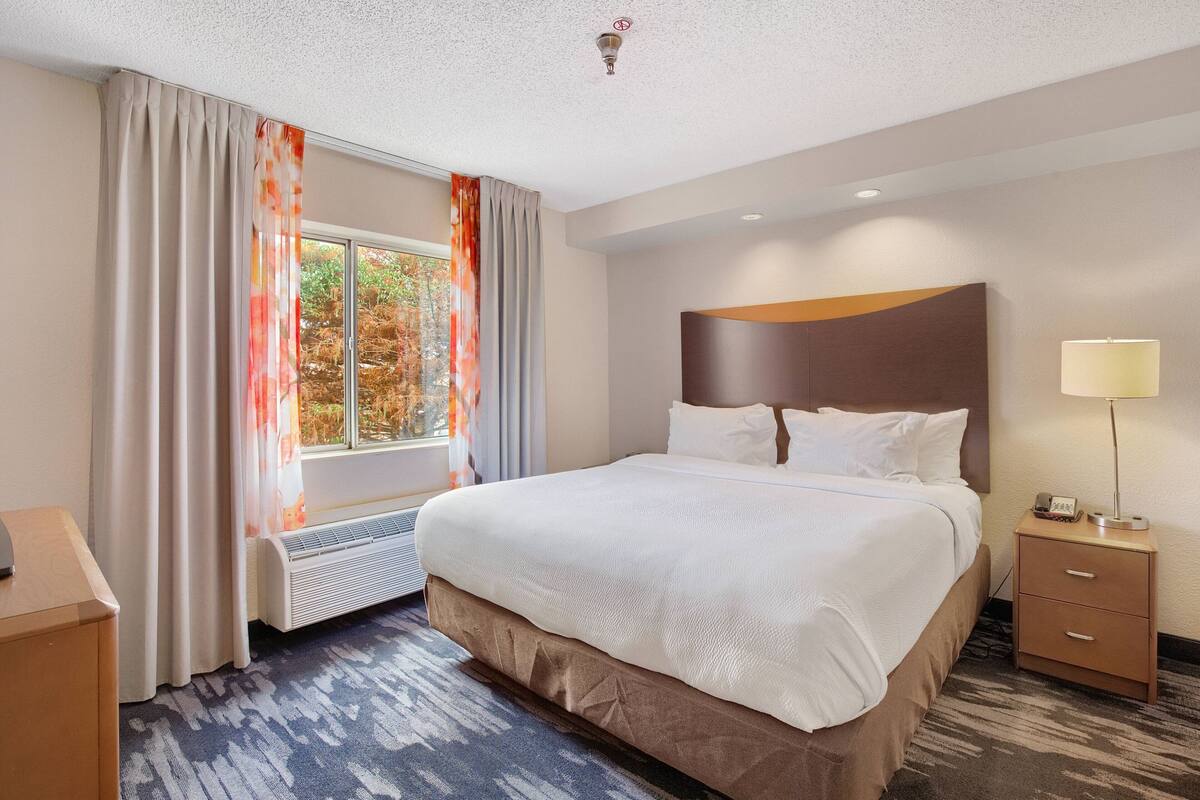 A hotel room with a large bed, a nightstand with a lamp, a window with curtains, and a wall-mounted heater.