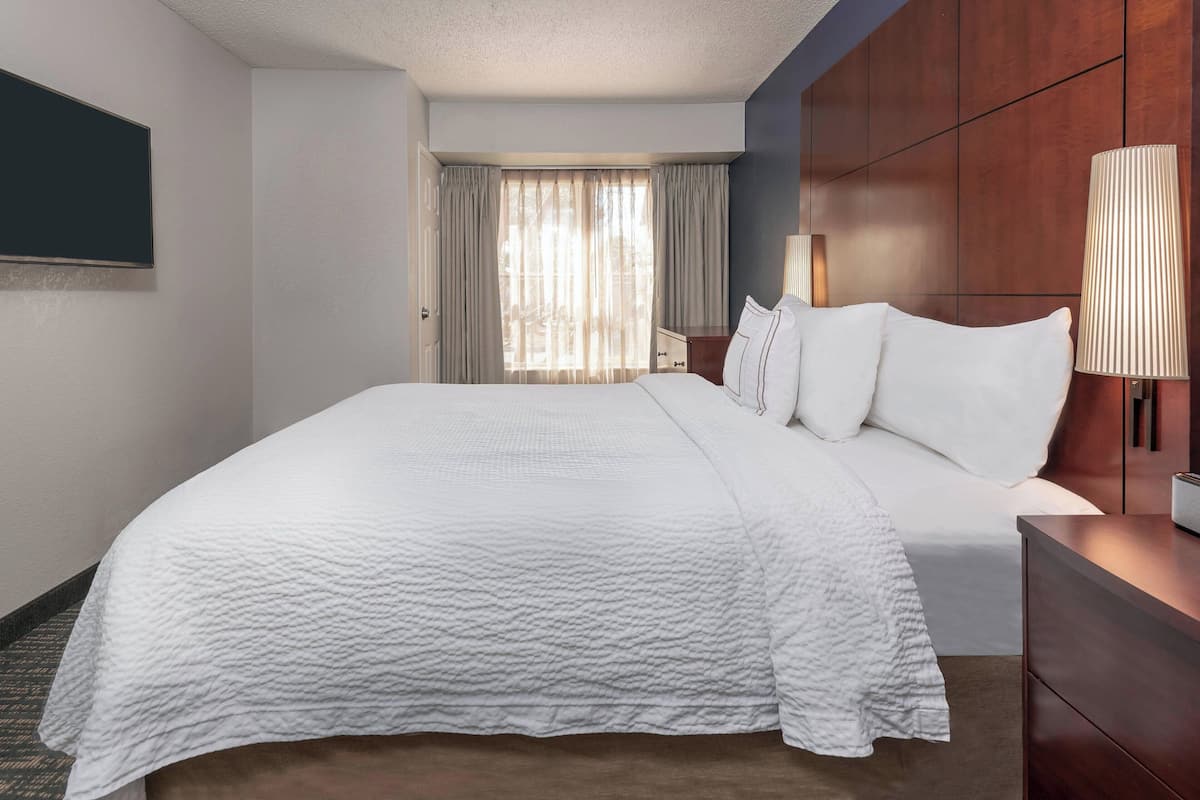 A hotel room with a large bed, a wooden wardrobe, a bedside lamp, and a view of the outdoors through sheer curtains.