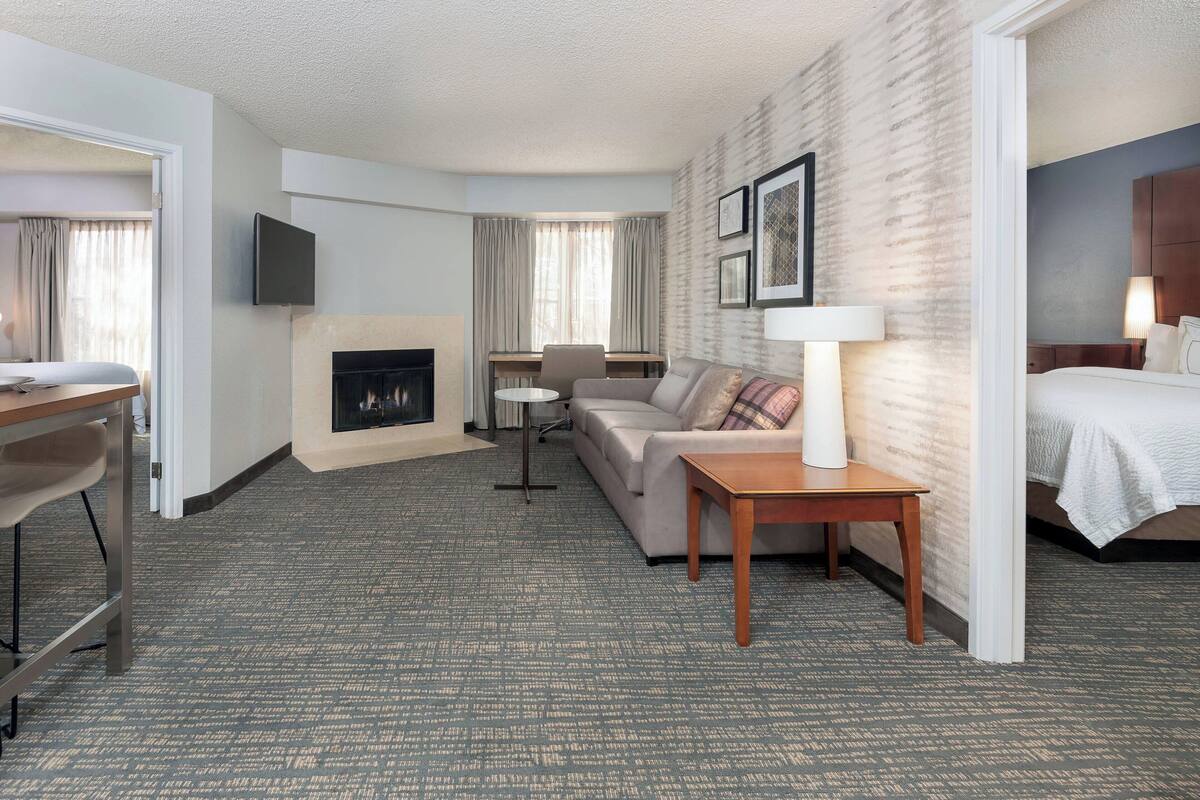 A hotel room with a sofa, a small table, a TV mounted on the wall, a fireplace, and a bed.