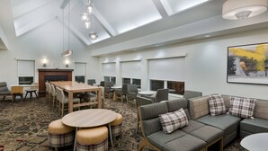 A modern hotel lobby with a high ceiling, skylights, a fireplace, a large sofa, and a dining area with round tables and chairs.