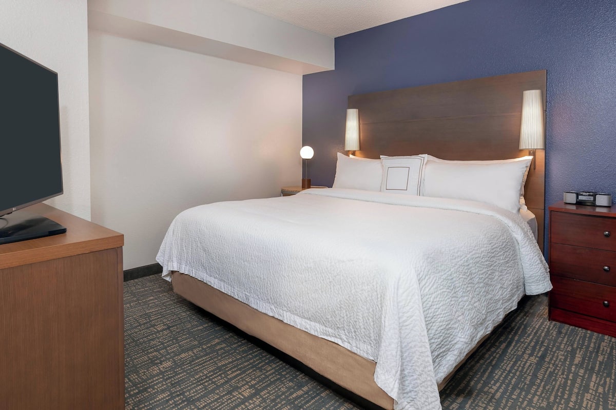 A hotel room with a large bed, a desk with a flat-screen TV, a nightstand with a lamp, and a dresser.