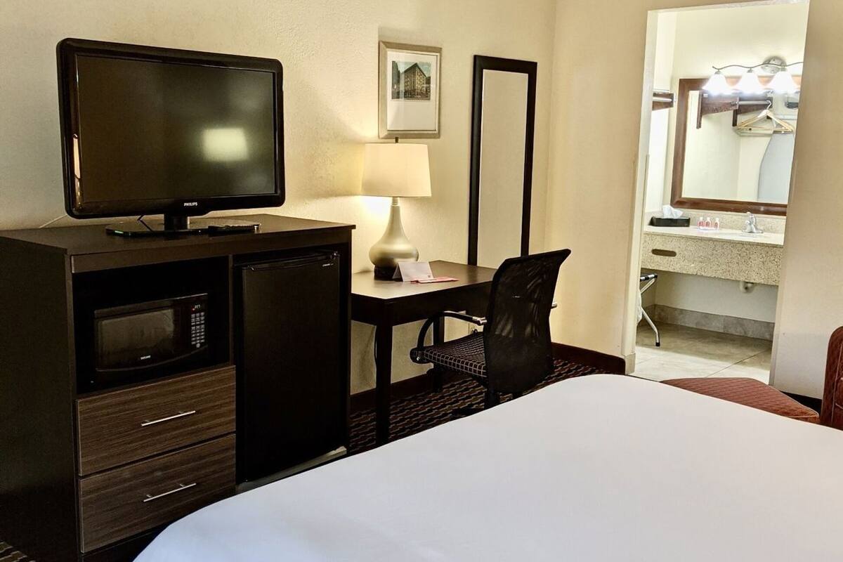 A hotel room with a bed, a desk with a television, a lamp, a chair, a dresser, and a bathroom mirror.