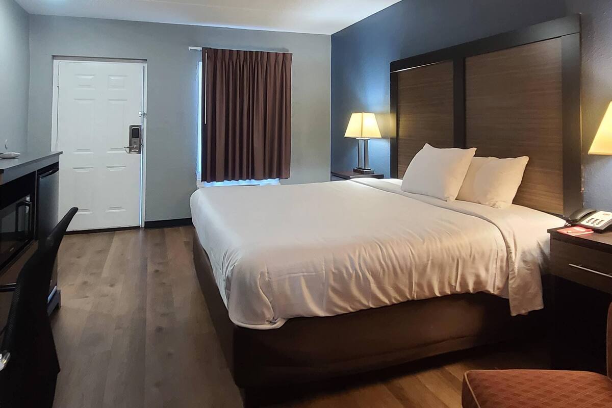 A hotel room with a large bed, a desk, a chair, a door, a window with curtains, and a bedside lamp.