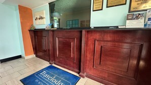 Un comptoir d’accueil avec un tapis bleu portant l’inscription « Travelodge By Windham », des certificats encadrés accrochés au mur et un tableau indiquant les heures d’arrivée et de départ.