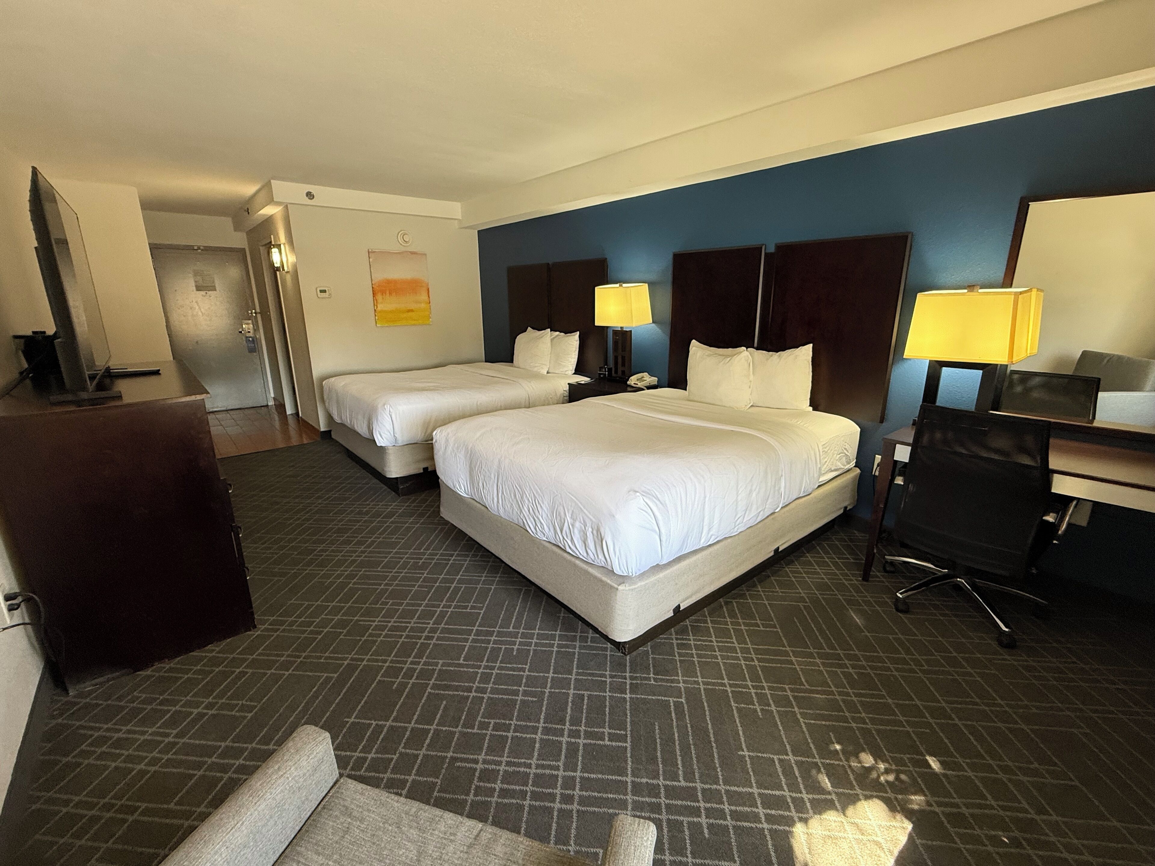 A hotel room with two beds, a desk, a chair, a lamp, and a small table.