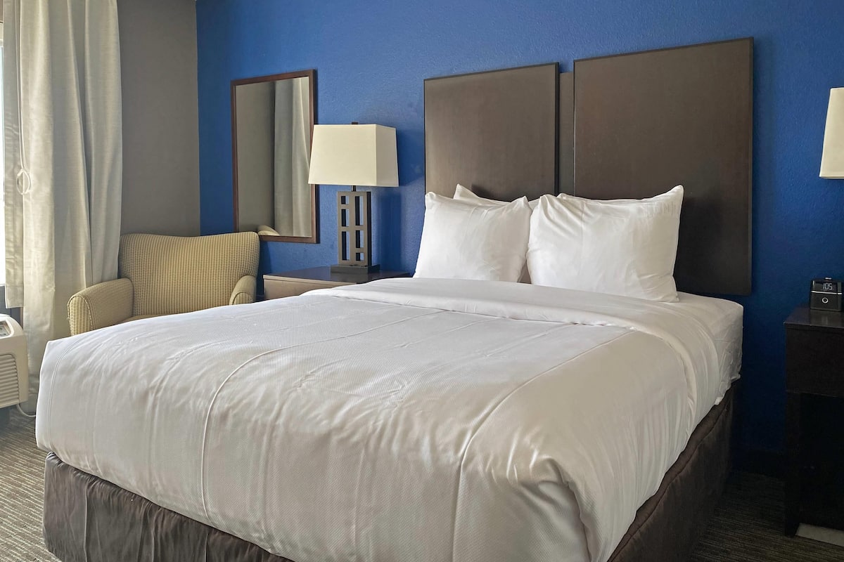A hotel room with a large bed, a nightstand with a lamp, a chair, a mirror, and a clock.