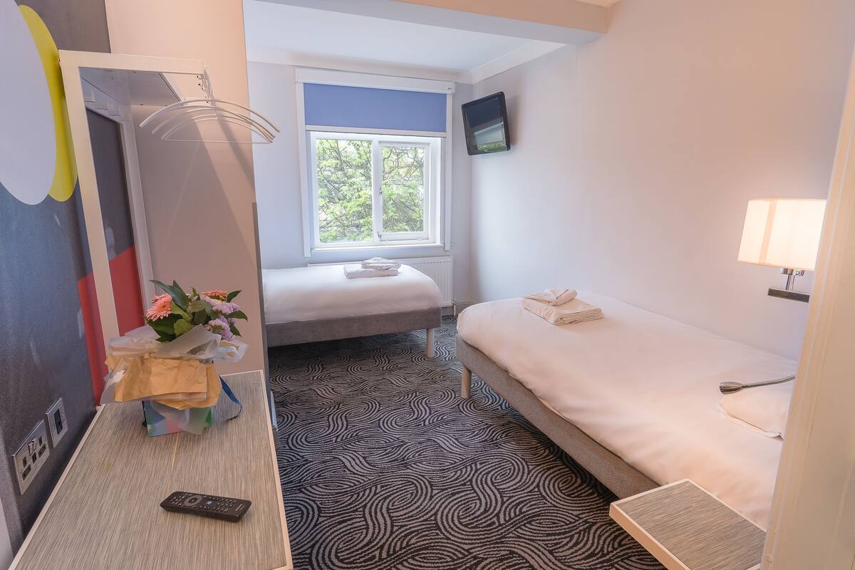 A hotel room with a bed, a bedside table with a remote control, a window with a view of trees, and a wall-mounted TV.