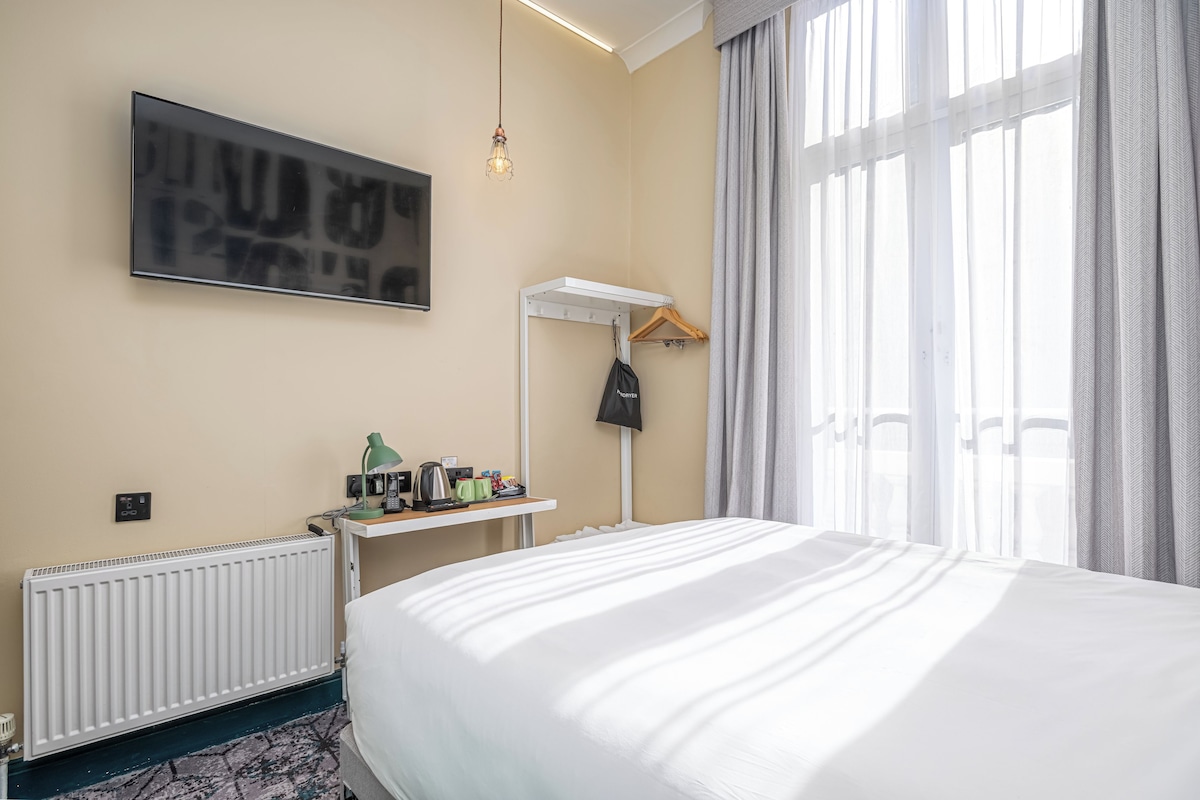 Quarto de hotel com cama, escrivaninha com abajur, radiador, cabideiro e janela com cortinas.