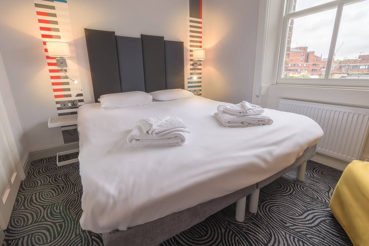 A neatly made bed with white linens and towels, a modern headboard, and a view of buildings through a window.