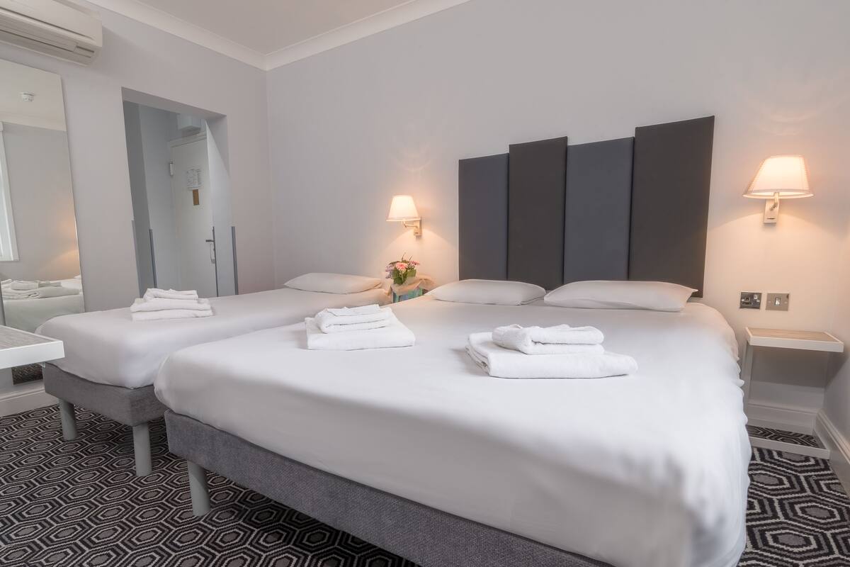 A hotel room with two beds, a large mirror, a small table, and a wall-mounted lamp.