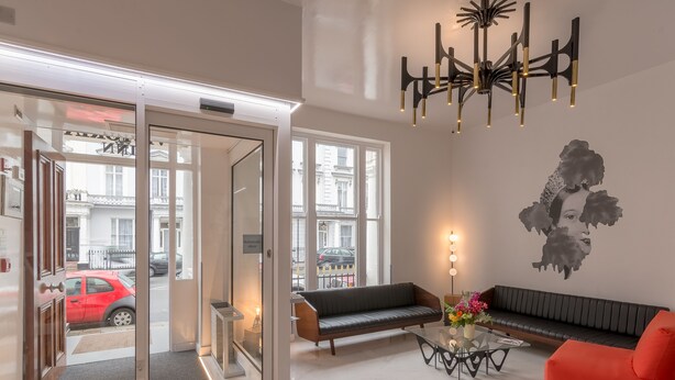 A modern lobby with a glass door entrance, a black sofa, a red armchair, a glass coffee table with flowers, and a wall-mounted abstract art piece. - Victoria Inn (London)