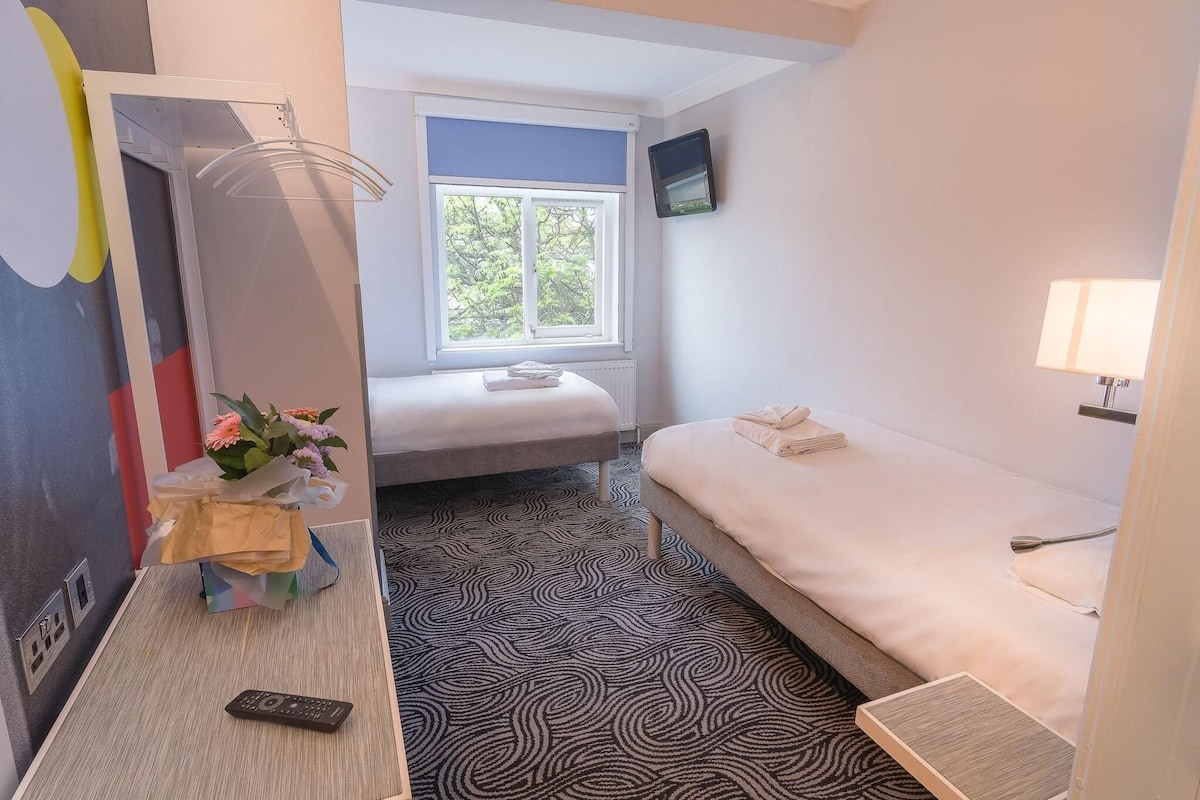 A compact hotel room with a bed, a small table with a remote control, a window with a view, and a wall-mounted TV.