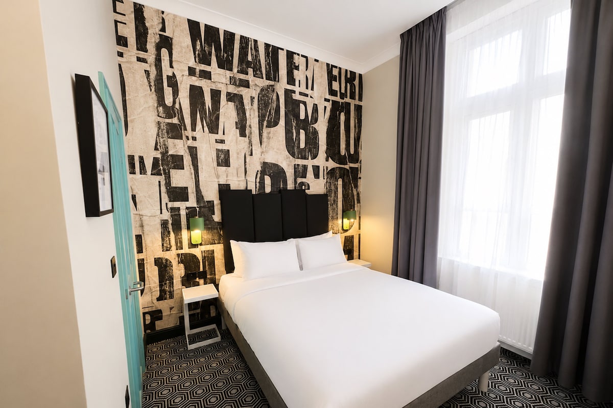 Quarto de hotel com uma cama grande, cabeceira com luminária e parede decorada com um design abstrato, além de uma janela com cortinas.
