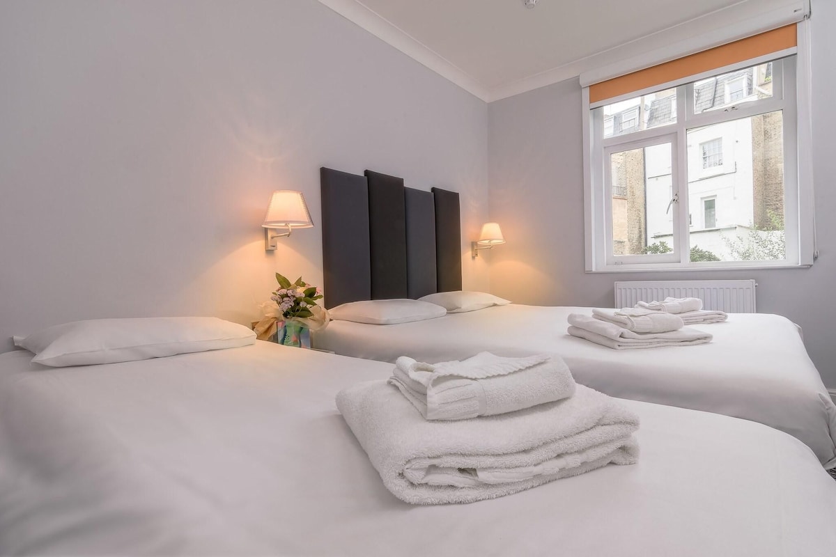 A hotel room with two beds, white linens, a large window with a view, and wall-mounted lamps.