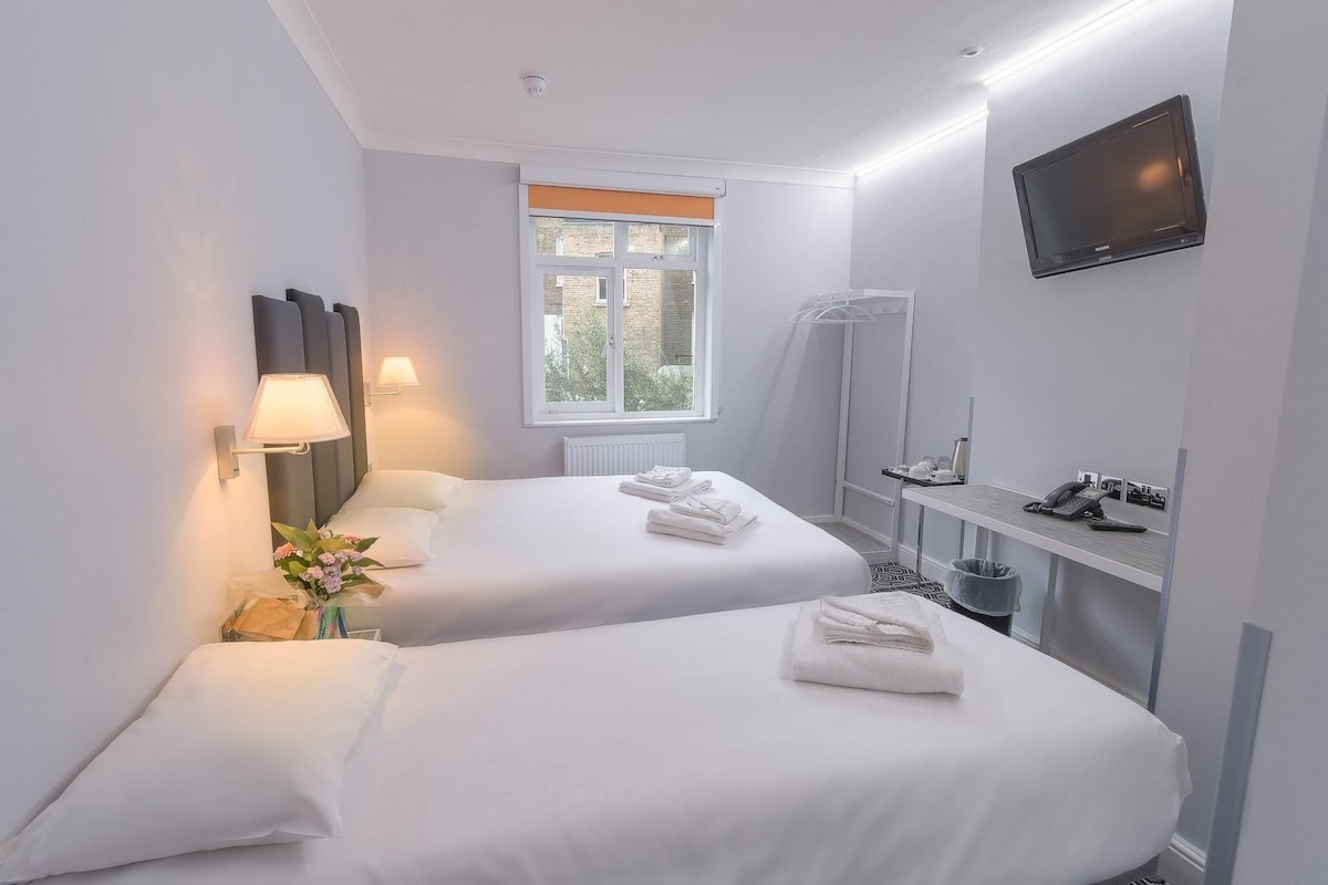 A hotel room with two beds, a TV, a window with blinds, a desk with a phone, and a trash bin.