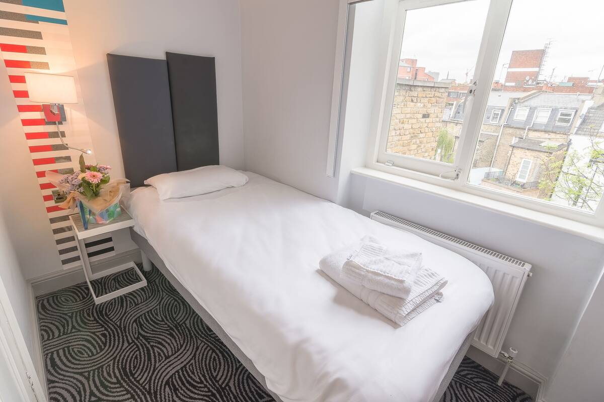 A small, neatly made bed with white linens and a folded towel, a bedside table with a lamp and a vase of flowers, and a window with a view of buildings.