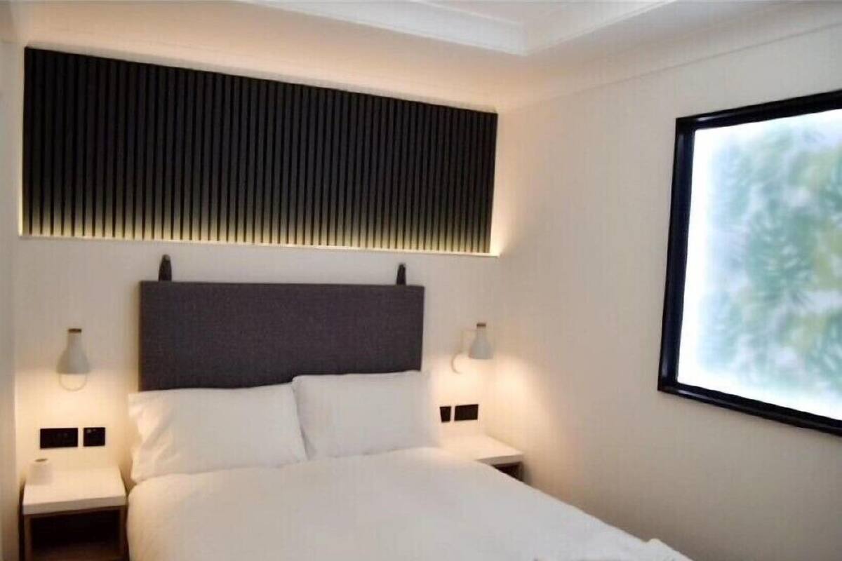 A modern hotel room with a large bed, two bedside tables, a headboard with vertical slats, and a large window with a view.