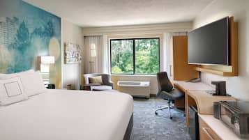 A hotel room with a large bed, a desk with a flat-screen TV, a chair, and a large window with a view of greenery.