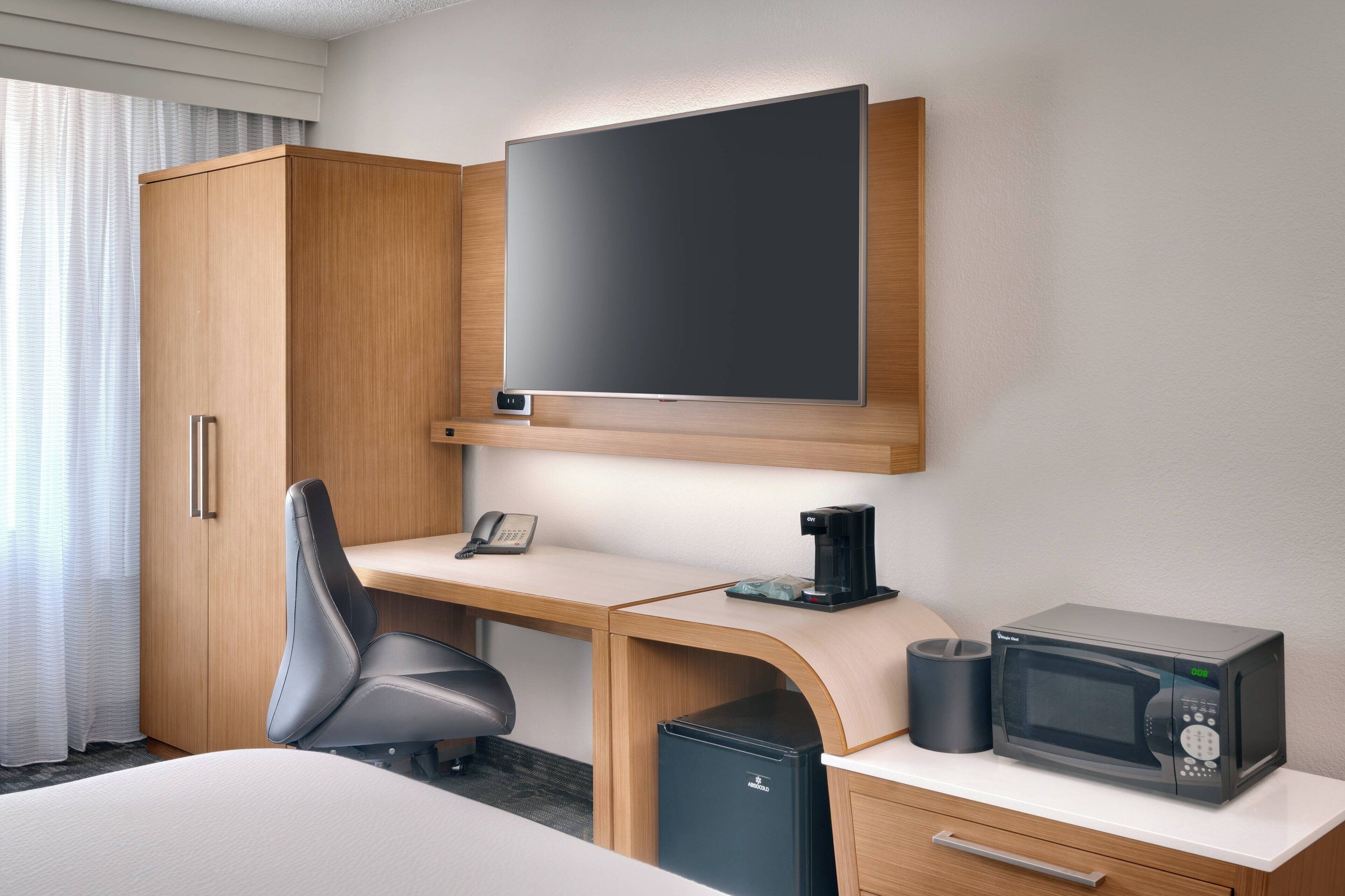 Ein Hotelzimmer mit einem Holztisch, einem schwarzen Bürostuhl, einem Flachbildfernseher, einer Kaffeemaschine, einer Mikrowelle und einem kleinen Mülleimer.