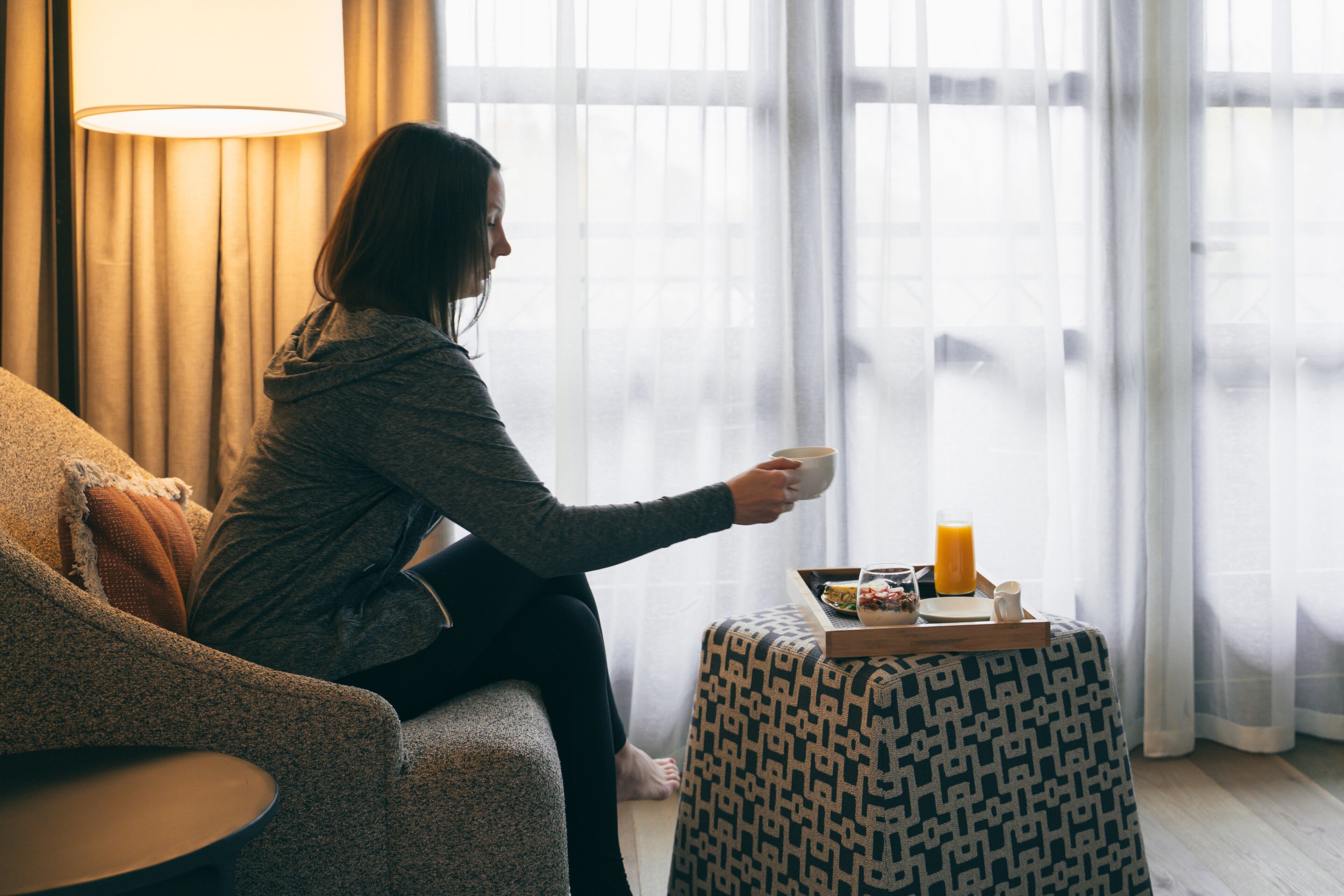 A person sitting on a sofa by a window with curtains, holding a cup. There is a tray with food and a glass of orange juice on a small table beside the sofa.
