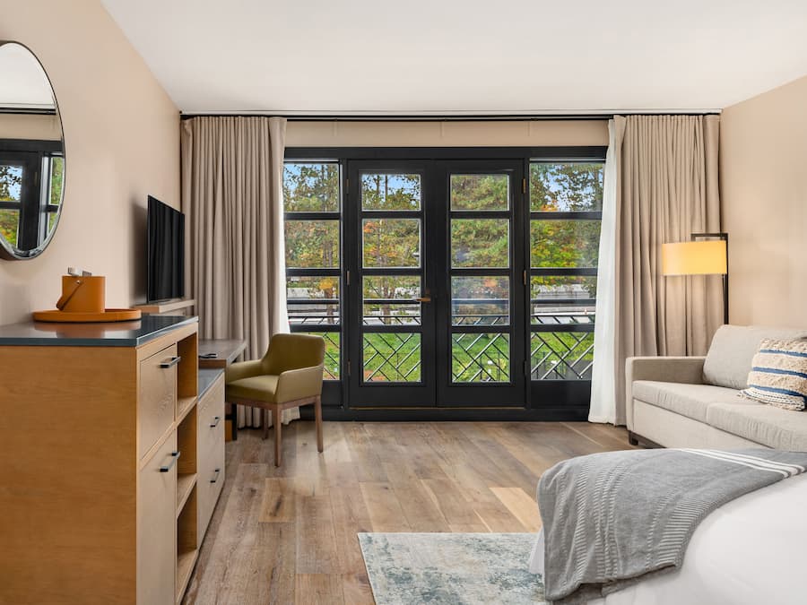Una habitación de hotel con un aparador de madera, un sofá, una cama, un escritorio con una silla, un televisor y ventanas grandes con cortinas.