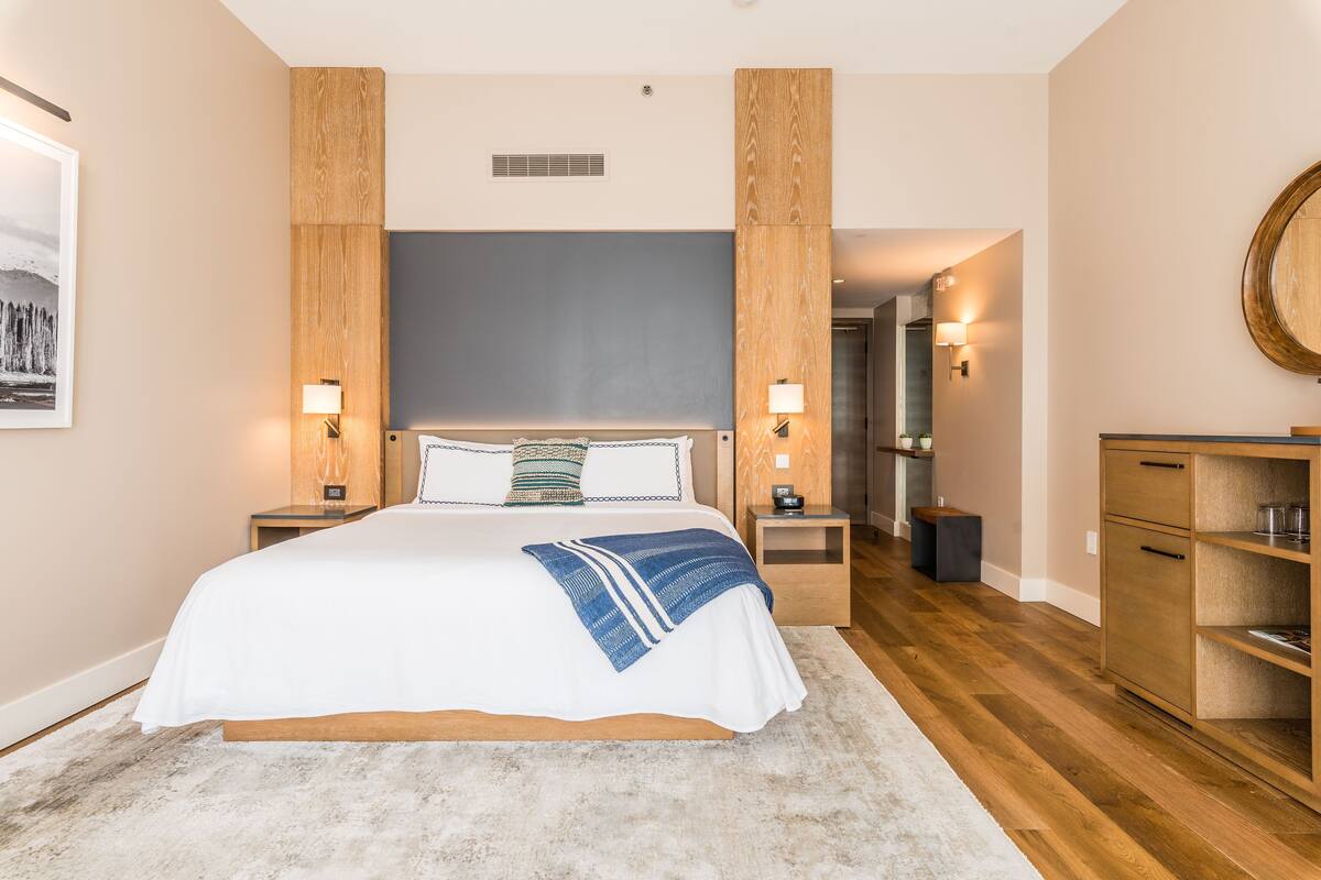 A modern hotel room with a large bed, wooden headboard, bedside lamps, a small nightstand, a wooden dresser, and a mirror.