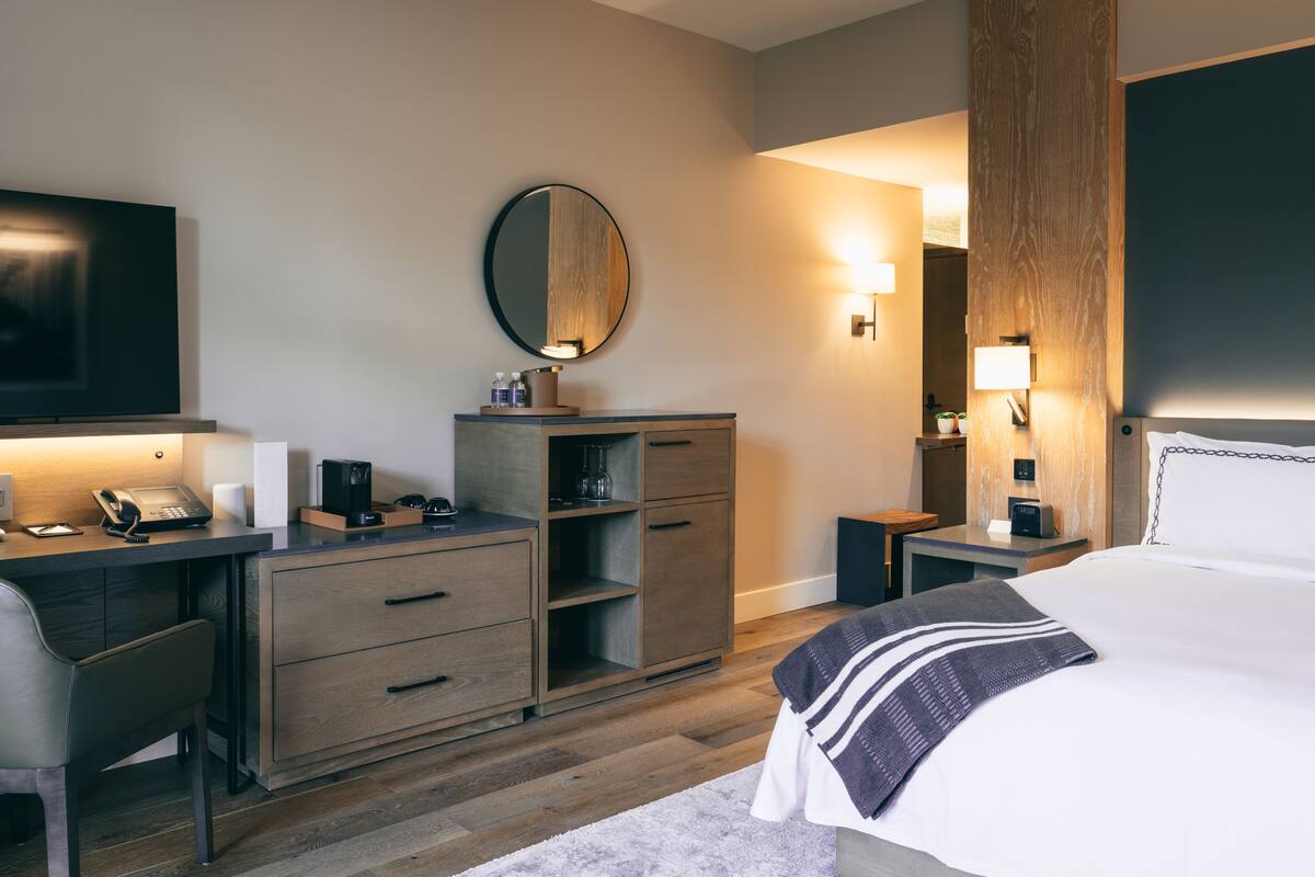 A hotel room with a desk, a bed, a dresser, a mirror, and a nightstand with a lamp.