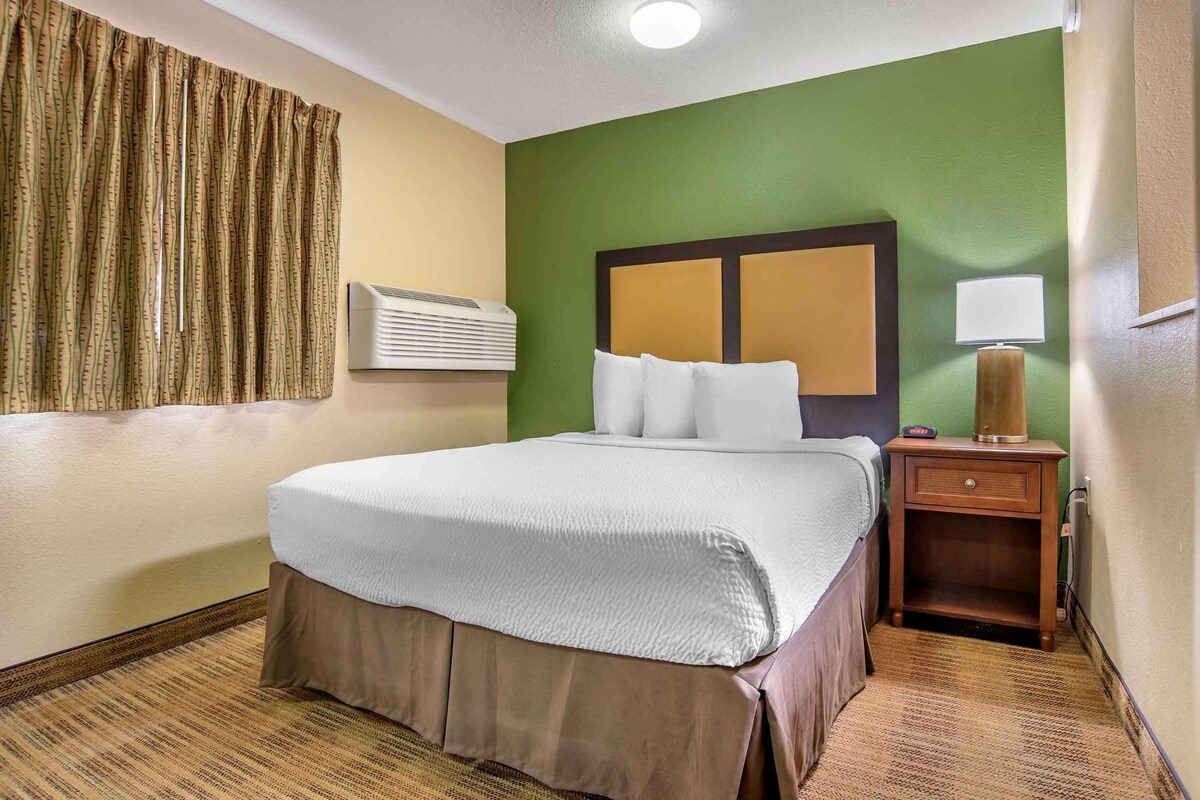 Habitación de hotel con cama, mesita de noche, lámpara, unidad de aire acondicionado y cortina.