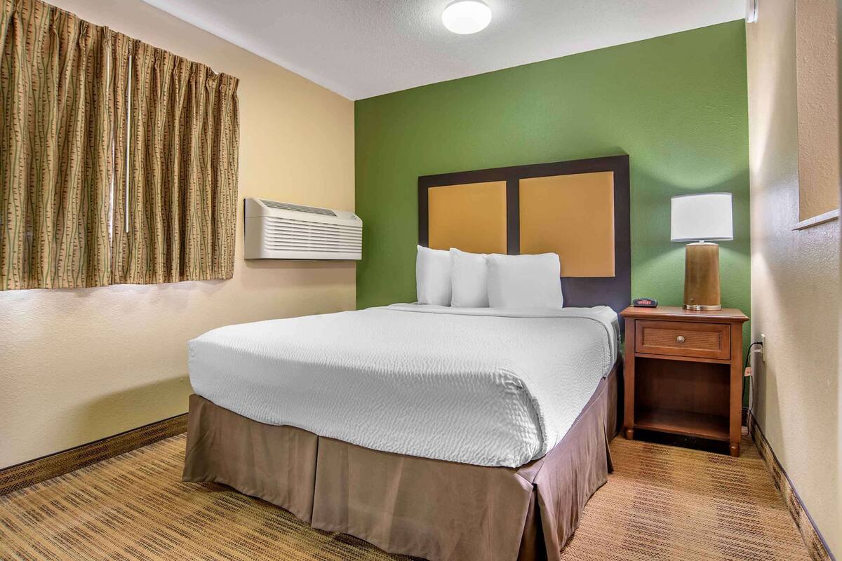 Ein Hotelzimmer mit Bett, Nachttisch, Lampe und Wandgerät zur Klimatisierung.