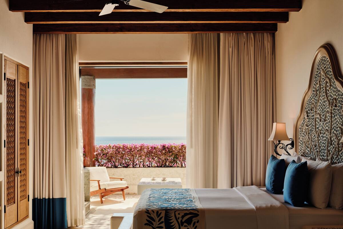 A hotel room with a bed, a nightstand, a lamp, a chair, and a view of the sea through large windows.