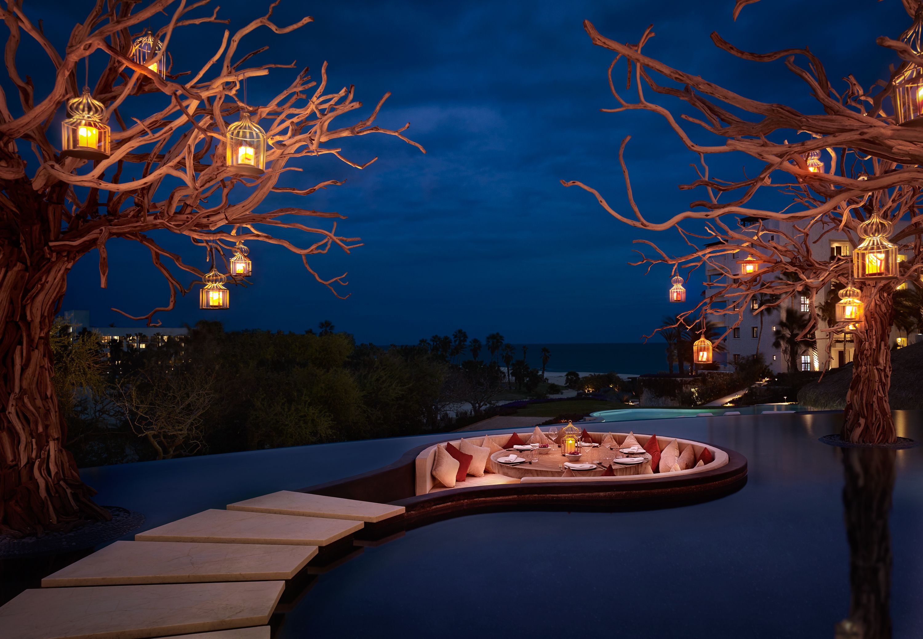 An outdoor dining area with a curved bench, lanterns, and a view of the ocean at night.